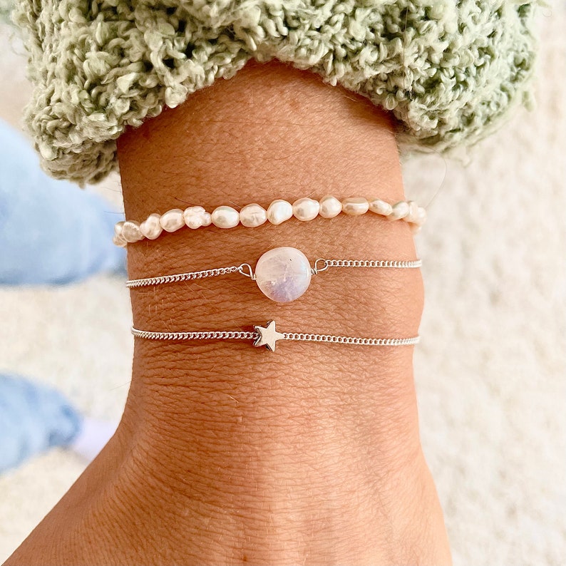 May include: Three bracelets stacked on a wrist. The top bracelet is a pearl strand. The middle bracelet is a silver chain with a large, round, white gemstone. The bottom bracelet is a silver chain with a star charm.