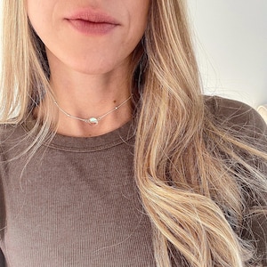 May include: A silver chain choker necklace with a small, round, iridescent pendant. The necklace is worn by a person with long, blonde hair and a brown shirt.