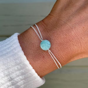 May include: A silver bracelet featuring a round, light blue gemstone. The bracelet has a delicate chain design and is worn on a wrist, paired with a white long-sleeved shirt. The gemstone is the focal point of the jewelry.