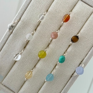 May include: A set of ten silver rings with different colored gemstone beads. The rings are displayed in a white fabric-lined box.