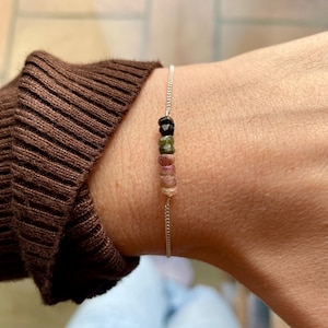 May include: A silver chain bracelet adorned with a line of multi-coloured tourmaline beads. The beads are in shades of black, green, and pink. The bracelet is displayed on a wrist, highlighting the jewellery's design and colour variations.
