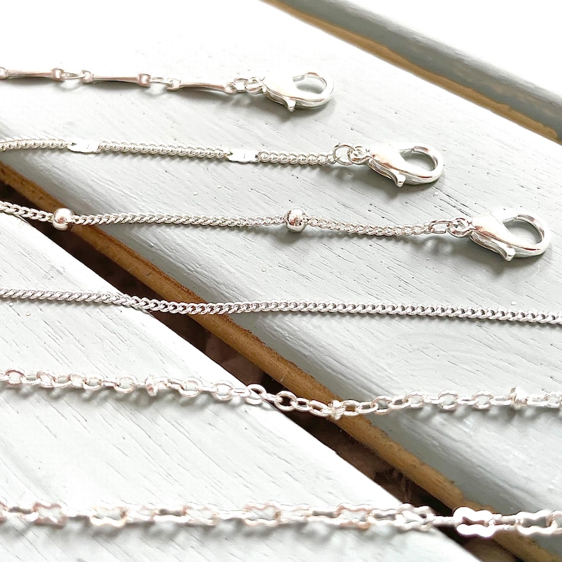 May include: Five silver chains of different styles and lengths, all with lobster claw clasps. The chains are arranged in a row on a light blue wooden surface.