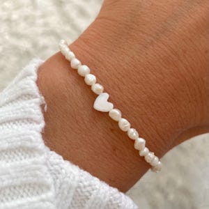 May include: A white pearl bracelet with a white heart charm. The bracelet is on a person's wrist.