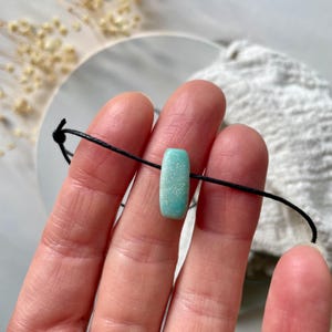 May include: A turquoise green gemstone bead on a black cord bracelet. The bead is rectangular and has a speckled texture.