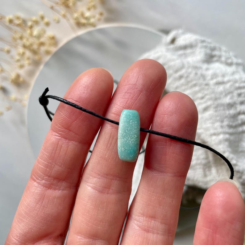May include: A turquoise green gemstone bead on a black cord bracelet. The bead is rectangular and has a speckled texture.