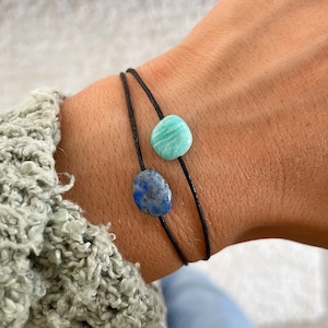 May include: Two bracelets with black string and round gemstone beads. One bead is a light blue lapis lazuli and the other is a light green aventurine.
