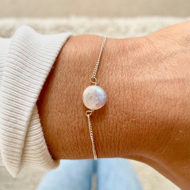 May include: A silver chain bracelet with a single round moonstone charm. The moonstone is iridescent and has a white and blue color.