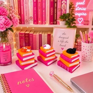 May include: Three stacks of miniature books in shades of orange and pink, each topped with a small cat figurine. The scene includes a pink spiral notebook with the word notes, a pink candle labeled focus inspire grow, and a framed sign with text.