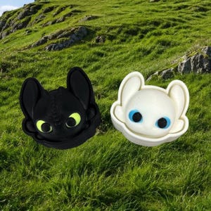 May include: Two small, molded plastic figures shaped like dragon heads. One figure is matte black with bright yellow eyes, and the other is matte white with vibrant blue eyes. Both figures feature large, rounded ears and a smooth, stylized design.