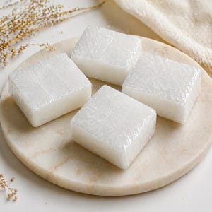 May include: Four square, white soap bars, individually wrapped in clear plastic, are arranged on a round, marble-like plate. The soap has a slightly translucent appearance. The plate is light-colored, and the background is a soft white.