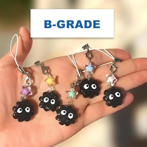 May include: A hand holds several black charm accessories. Each charm features a round, fluffy character with white eyes, and is adorned with a star-shaped bead in various colors. The text "B-GRADE" is displayed in a white rectangle.