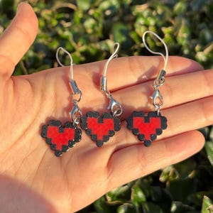 May include: Three pixelated heart charms in red and black, each with a silver clasp and white cord. The charms are held in a hand against a backdrop of green foliage. The heart design is reminiscent of retro video game graphics.
