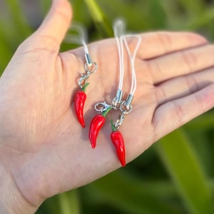 May include: Three red chili pepper charms with green stems and silver-colored metal hardware. The charms are attached to white cords. The background is blurred green foliage.