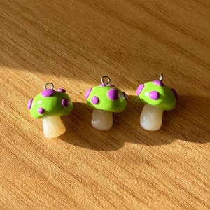 Op de afbeelding: Drie handgemaakte paddenstoelbedels. Elke bedel heeft een groene hoed met paarse stippen, een witte steel en een zilveren lus om aan sieraden of andere knutselwerken te bevestigen. De bedels worden op een houten oppervlak gepresenteerd.