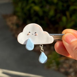 Op de afbeelding: Een witte, wolk-vormige haarclip met een lachend gezicht en twee lichtblauwe tranen die eronder hangen. Het haaraccessoire wordt door een hand vastgehouden tegen een vage groene achtergrond.