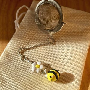 May include: A stainless steel tea infuser with a chain, a white daisy charm, and a yellow and black bee charm. The infuser sits on a beige linen bag, perfect for steeping loose leaf tea.