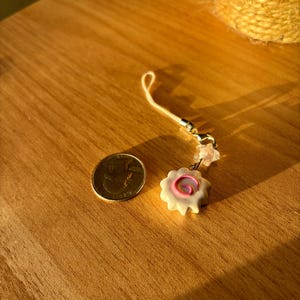 Peut inclure: Un petit porte-bonheur blanc avec un motif en spirale rose, &agrave; c&ocirc;t&eacute; d'une pi&egrave;ce de monnaie. Le porte-bonheur est attach&eacute; &agrave; une cha&icirc;ne dor&eacute;e avec une boucle. Les objets sont sur une surface en bois.