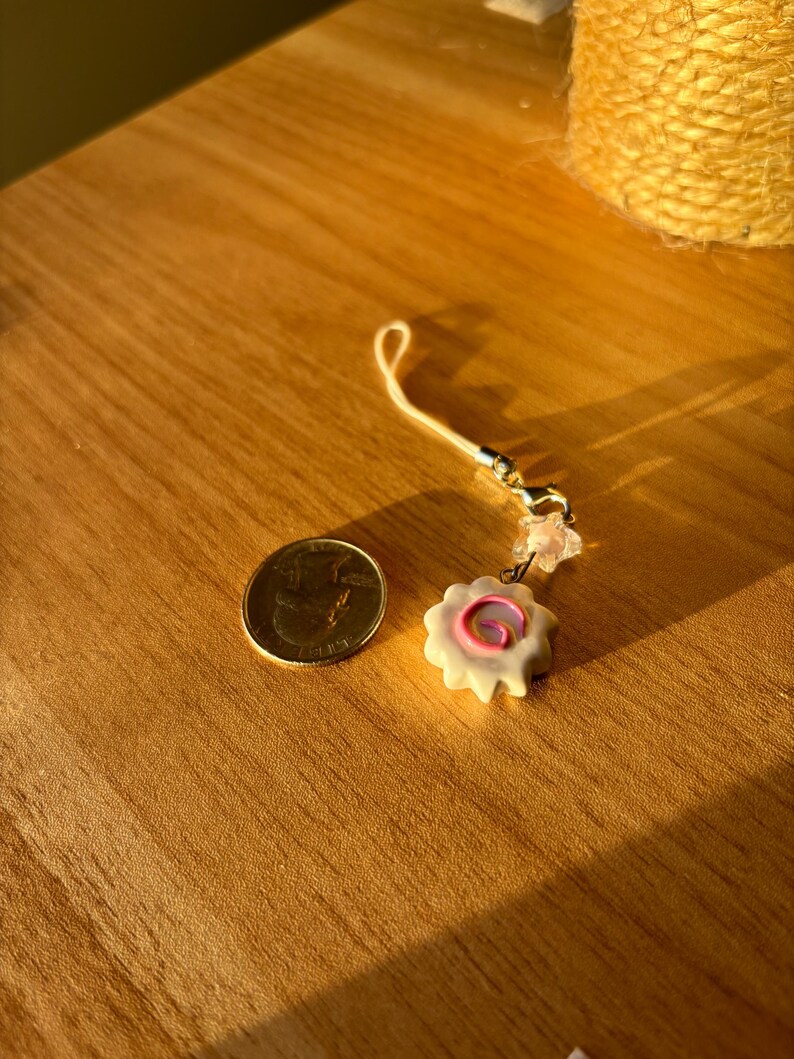 Peut inclure: Un petit porte-bonheur blanc avec un motif en spirale rose, &agrave; c&ocirc;t&eacute; d'une pi&egrave;ce de monnaie. Le porte-bonheur est attach&eacute; &agrave; une cha&icirc;ne dor&eacute;e avec une boucle. Les objets sont sur une surface en bois.