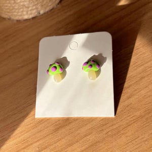 Op de afbeelding: Een paar paddenstoelvormige oorstekers. De oorbellen hebben een felgroene dop met roze stippen en een crèmekleurige steel. Ze worden gepresenteerd op een witte oorbellenkaart, ideaal om cadeau te geven of tentoon te stellen.