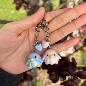 Puede incluir: Un llavero con dos pequeños colgantes de pingüino, uno azul y otro rosa, con un pequeño colgante de corazón en el medio. Los colgantes están unidos a una cadena plateada.