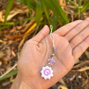 Peut inclure: Un charm narutomaki blanc et rose avec un motif en spirale rose, orn&eacute; d'une perle en forme d'&eacute;toile violette. Le charm est attach&eacute; &agrave; un fermoir argent&eacute; et &agrave; une ficelle blanche, parfait pour un accessoire de t&eacute;l&eacute;phone ou de sac.