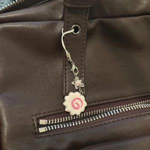 Peut inclure: Un sac en cuir marron avec une fermeture &eacute;clair argent&eacute;e et un charm d&eacute;coratif. Le charm pr&eacute;sente un motif tourbillon blanc et rose, une petite &eacute;toile et une perle transparente. Le charm est attach&eacute; au sac avec une cha&icirc;ne argent&eacute;e.