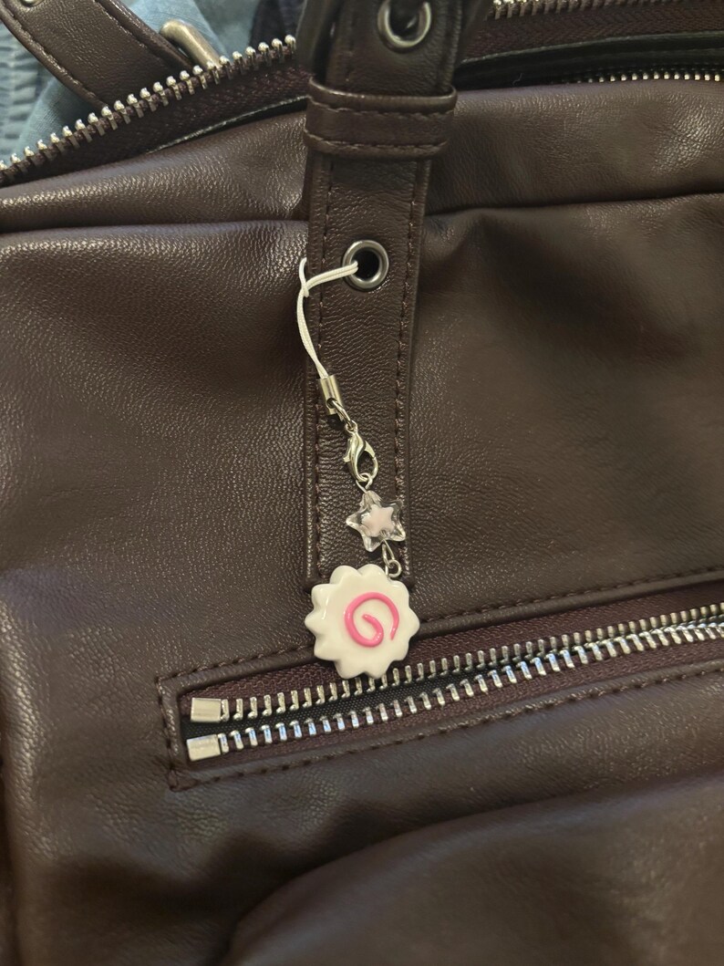 Peut inclure: Un sac en cuir marron avec une fermeture &eacute;clair argent&eacute;e et un charm d&eacute;coratif. Le charm pr&eacute;sente un motif tourbillon blanc et rose, une petite &eacute;toile et une perle transparente. Le charm est attach&eacute; au sac avec une cha&icirc;ne argent&eacute;e.