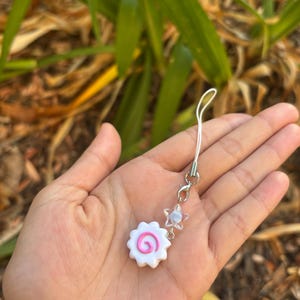 Peut inclure: Un charm blanc et rose avec un motif en spirale, ressemblant &agrave; un narutomaki. Il est attach&eacute; &agrave; une cha&icirc;ne argent&eacute;e avec une perle en forme d'&eacute;toile. Le charm est tenu dans une main sur un fond flou de feuillage vert.