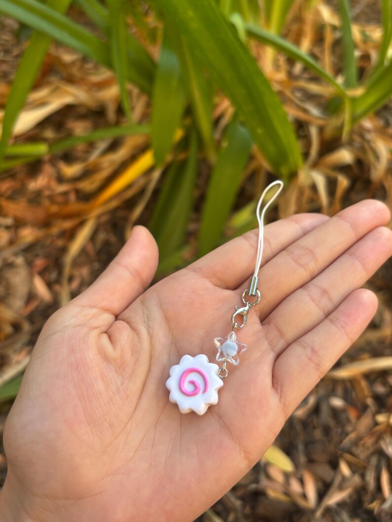 Peut inclure: Un charm blanc et rose avec un motif en spirale, ressemblant &agrave; un narutomaki. Il est attach&eacute; &agrave; une cha&icirc;ne argent&eacute;e avec une perle en forme d'&eacute;toile. Le charm est tenu dans une main sur un fond flou de feuillage vert.