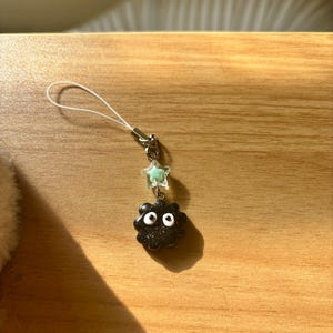 Może przedstawiać: Czarny charms z białymi oczami i charms w kształcie gwiazdy. Charms jest przymocowany do srebrnego metalowego zapięcia i białego sznurka. Charms znajduje się na drewnianej powierzchni.