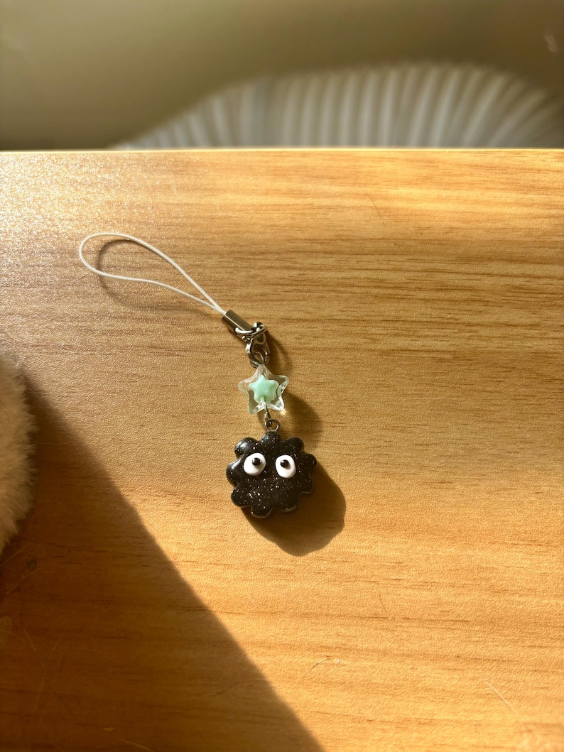 Może przedstawiać: Czarny charms z białymi oczami i charms w kształcie gwiazdy. Charms jest przymocowany do srebrnego metalowego zapięcia i białego sznurka. Charms znajduje się na drewnianej powierzchni.