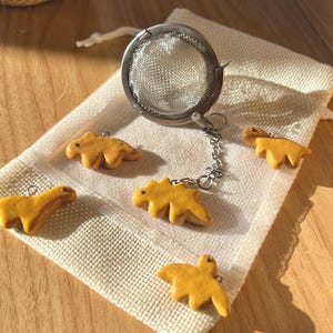 May include: A stainless steel tea infuser with a chain and small, yellow dinosaur-shaped charms. The infuser sits on a small, off-white linen bag, with several dinosaur charms scattered around it. The background is a light-colored wooden surface.