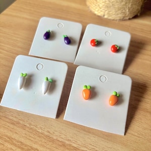 May include: Four pairs of stud earrings, each featuring a different vegetable design. The earrings include eggplants, tomatoes, carrots, and radishes, all with detailed, colorful designs. Each pair is mounted on a white earring card.
