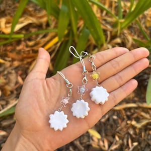 Peut inclure: Trois breloques de t&eacute;l&eacute;phone faites &agrave; la main avec des pendentifs blancs en forme de fleur. Chaque breloque a une perle en forme d'&eacute;toile de couleur diff&eacute;rente et une cha&icirc;ne argent&eacute;e. Les breloques sont tenues dans une main sur un fond de feuillage vert.