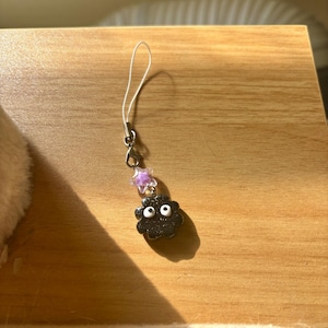Może przedstawiać: Czarny charms z białymi oczami i charms w kształcie gwiazdy. Charms jest przymocowany do srebrnego metalowego zapięcia i białego sznurka. Charms znajduje się na drewnianej powierzchni.