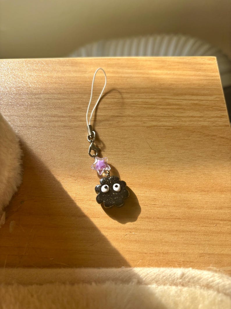 Może przedstawiać: Czarny charms z białymi oczami i charms w kształcie gwiazdy. Charms jest przymocowany do srebrnego metalowego zapięcia i białego sznurka. Charms znajduje się na drewnianej powierzchni.