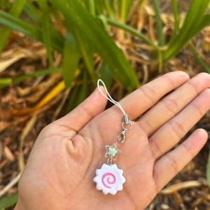 Peut inclure: Un charm blanc et rose avec un motif en spirale, attach&eacute; &agrave; une cha&icirc;ne argent&eacute;e avec deux breloques en forme d'&eacute;toile. Le charm est tenu dans une main sur un fond de feuillage vert.