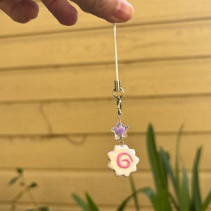 Peut inclure: Un charm blanc et rose avec un motif en spirale, suspendu &agrave; une cha&icirc;ne argent&eacute;e. Au-dessus, une petite &eacute;toile violette translucide. Le charm est attach&eacute; &agrave; un cordon blanc fin.
