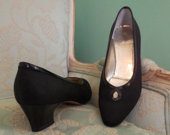 Puede incluir: Un par de zapatos negros de tacón alto con punta puntiaguda y un pequeño detalle decorativo en la parte delantera. Los zapatos están sobre una silla de color azul claro con estampado.
