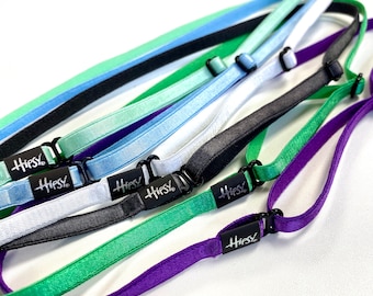 May include: A collection of adjustable elastic headbands in various colors including light green, light blue, white, black, and purple. Each headband features a black plastic slider for size adjustment and a black tag with the white text Hipsy.