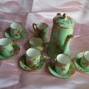 Puede incluir: Un juego de té de porcelana verde menta que incluye una tetera con tapa, una lechera, un azucarero y cuatro tazas con sus platos a juego. Cada pieza está decorada con bordes dorados y un estampado floral en tonos rosa, blanco y marrón.