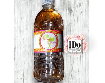May include: A clear plastic water bottle with a label that says "We Do" with a palm tree and stars. The label also says "Whitney & Justin" and has a pink and orange color scheme. The bottle is sitting on a wooden surface with a pink flower in the background. The label also includes the text "I Do Artsy Weddings, A Custom Label & Paper Goods Boutique."