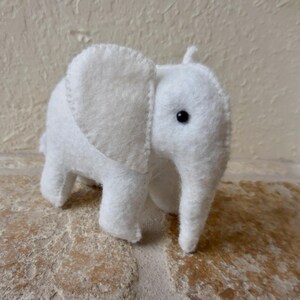 May include: A white felt elephant toy with black eyes. The elephant is standing on a light brown surface.