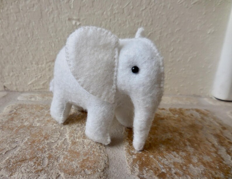 May include: A white felt elephant toy with black eyes. The elephant is standing on a light brown surface.