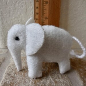 May include: A small white felt elephant toy with black eyes. The elephant has a long trunk and a curled tail.