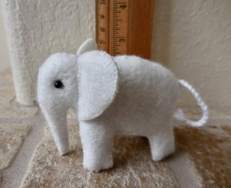 May include: A small white felt elephant toy with black eyes. The elephant has a long trunk and a curled tail.