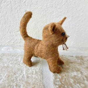 May include: A small, brown, needle-felted cat figurine standing on four legs with its tail held upright. The cat has dark, round eyes, pointed ears, and thin, light-colored whiskers protruding from its snout, crafted from textured wool fibers.