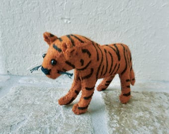 May include: A handmade orange felt tiger figurine with black stripes. The tiger has small black eyes and a dark blue nose. It is standing on a light-colored stone surface. The tiger is a cute, handcrafted animal figure.