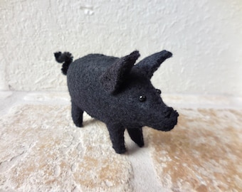May include: A small, handcrafted black pig figurine made of felted wool. The pig features a textured, fuzzy surface, two pointed ears, a curled tail, and two small, round, black bead eyes set into the head. The snout is slightly raised.