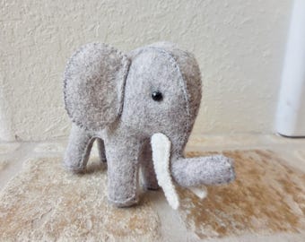 May include: A small, handcrafted felt elephant figurine in a light gray color. The plush animal features a rounded body, large ears, a trunk, and two white felt tusks. A single black bead serves as the eye, and visible stitching outlines the edges.